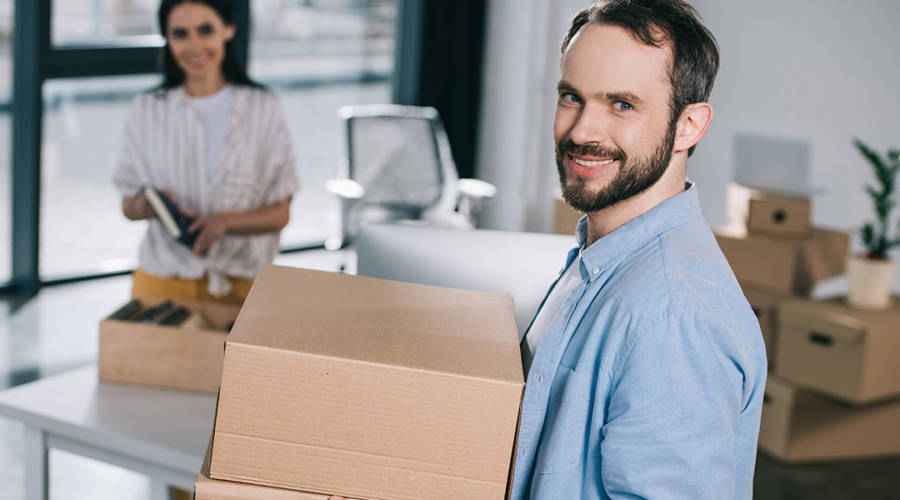 A man in light blue shirt moving boxes into a new office space with female coworker unpacking boxes in the background