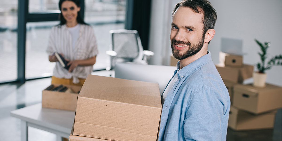 A man in light blue shirt moving boxes into a new office space with female coworker unpacking boxes in the background